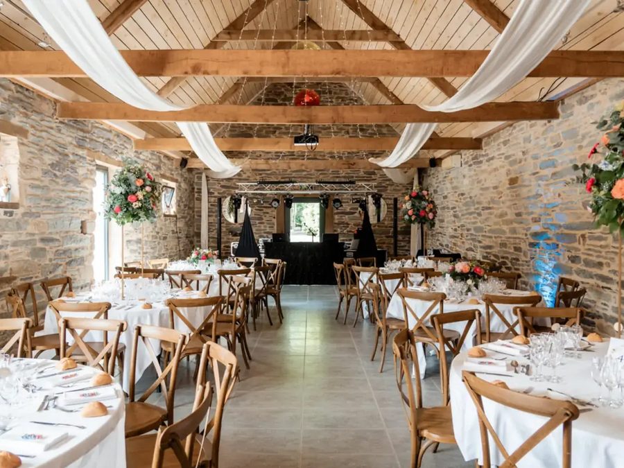 La salle de réception : murs de pierre, poutres en chêne et voilages blancs au-dessus des tables rondes
